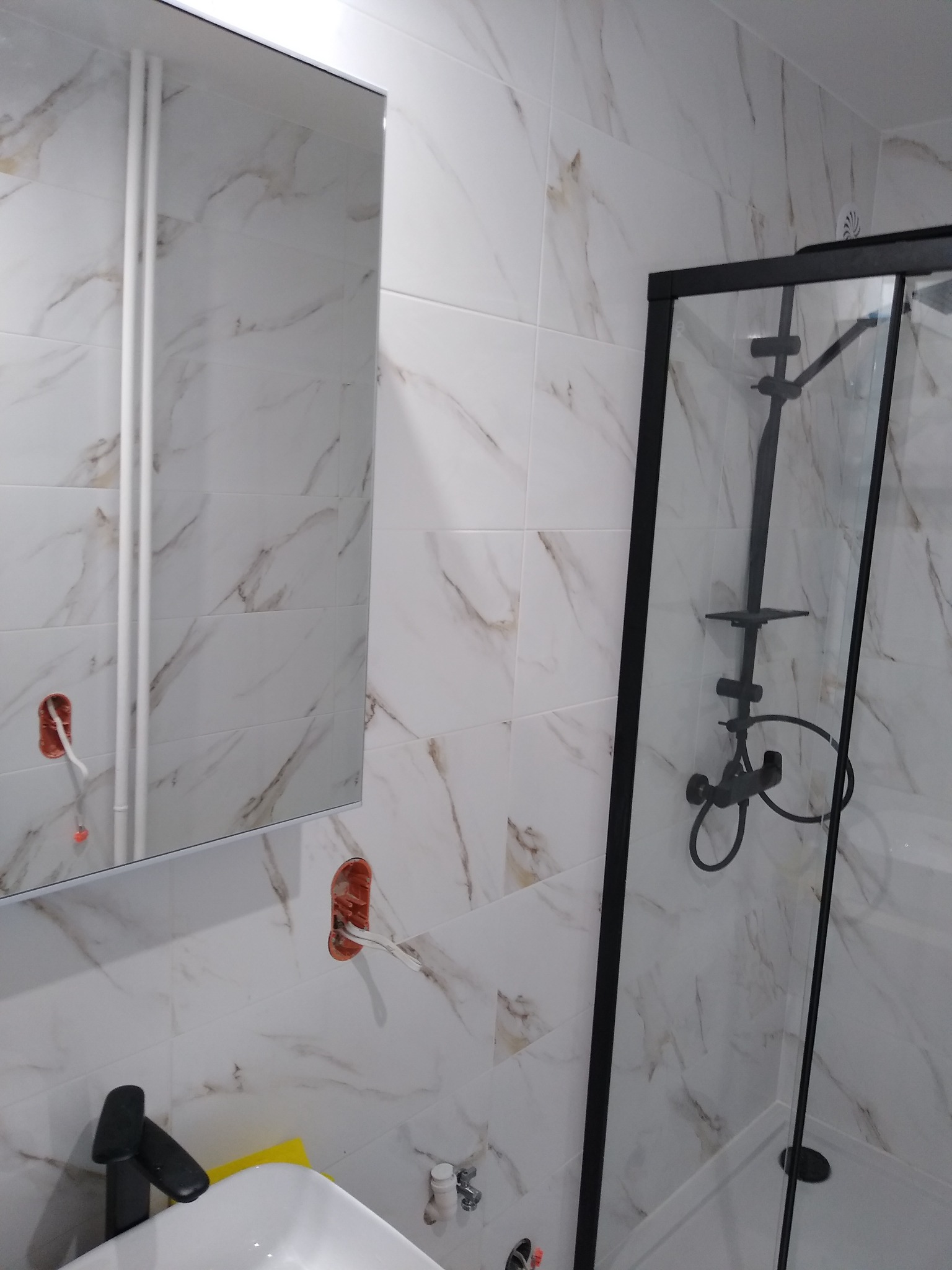 Białe marmurowe płytki w łazience z czarną armaturą, kabiną prysznicową i lustrem z widocznymi elementami instalacji elektrycznej.