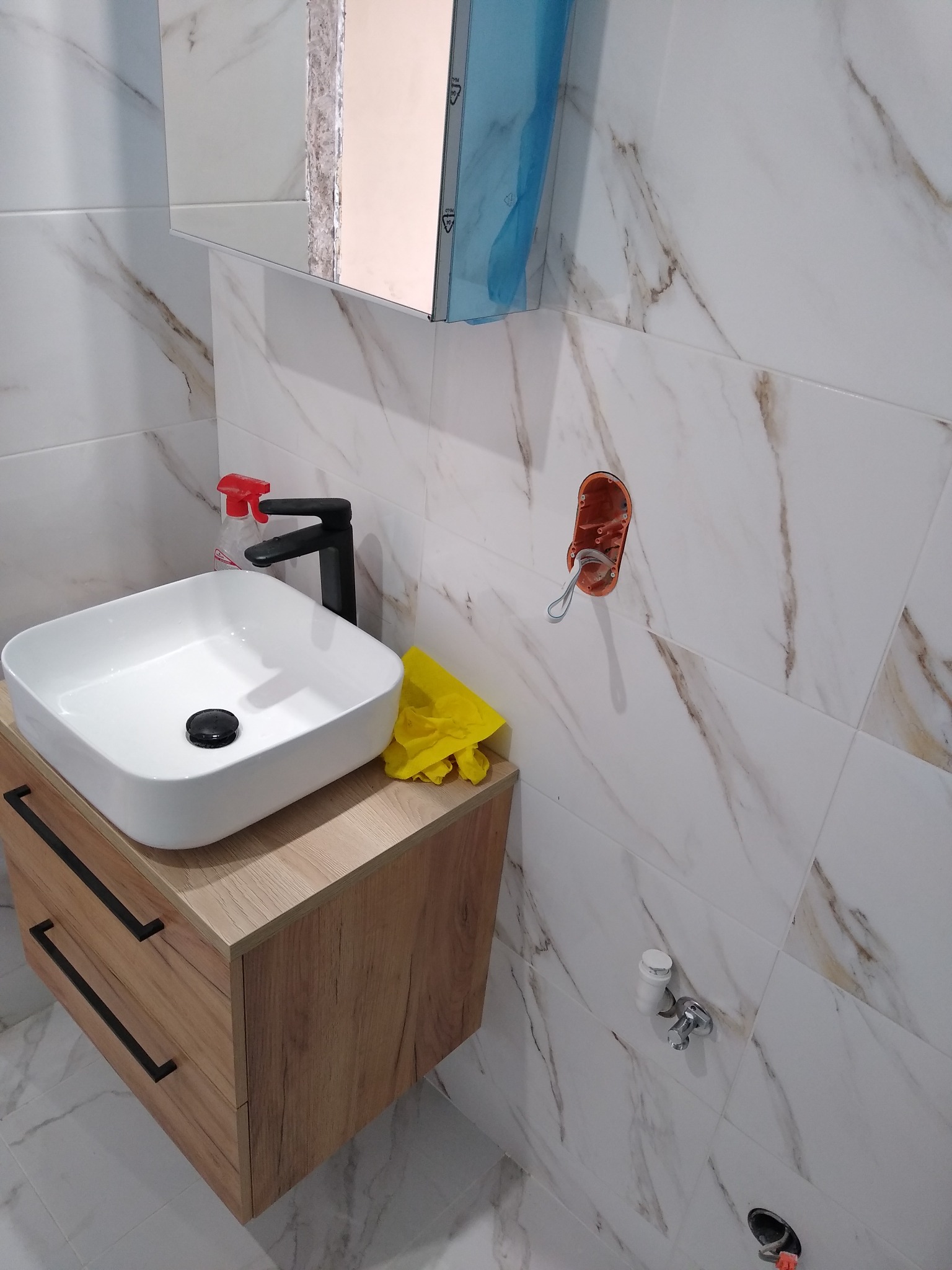 Nowoczesna umywalka nablatowa w kształcie kwadratu z czarną armaturą, zamontowana na szafce z drewnopodobnym frontem, białe płytki imitujące marmur na ścianie, widoczna puszka elektryczna bez...