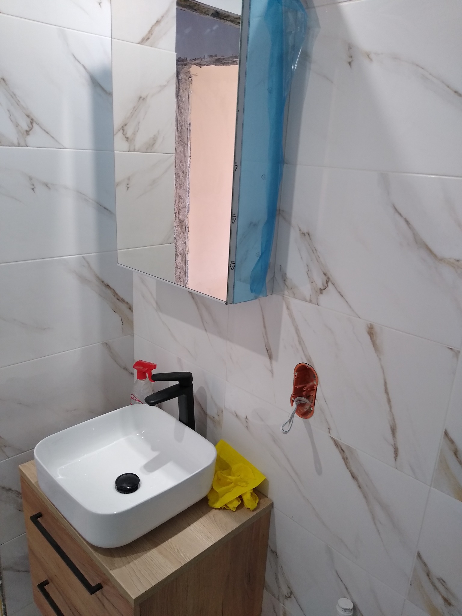 Biała, kwadratowa umywalka nablatowa z czarną armaturą i drewnopodobną szafką, marmurowe płytki ścienne, częściowo zamontowane lustro z folią ochronną, widoczna puszka elektryczna i butelka środka...