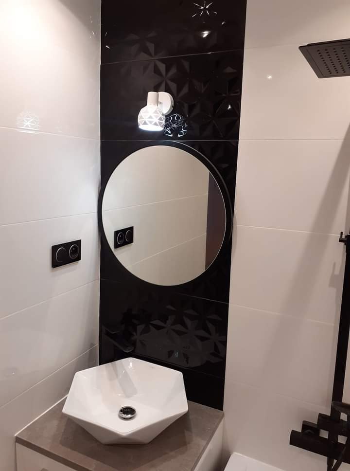 Nowoczesna łazienka z biało-czarnymi płytkami, okrągłym lustrem, sześciokątną umywalką nablatową i czarną armaturą prysznicową.