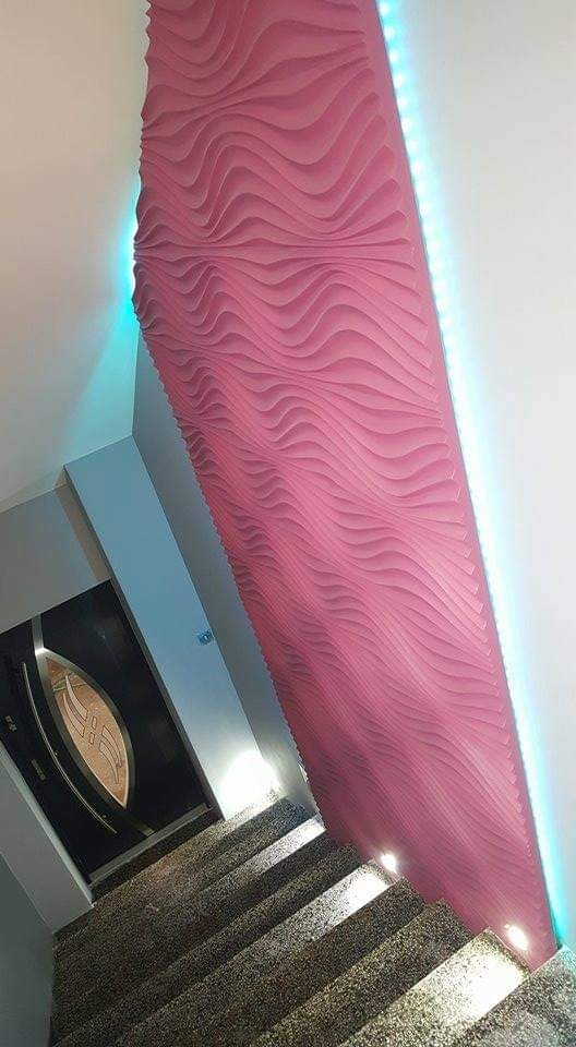 Różowa, falista dekoracja ścienna z podświetleniem LED wzdłuż krawędzi, widok z dołu schodów z kamiennymi stopniami i czarnymi drzwiami z elementami dekoracyjnymi.