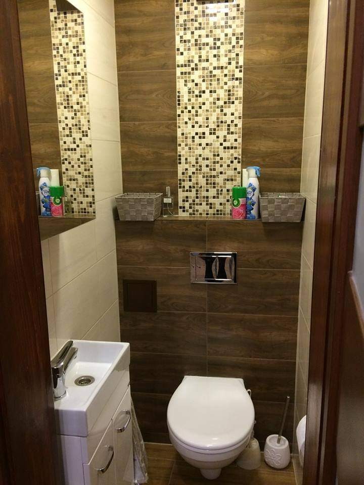 Mała toaleta z umywalką, wykończona brązowymi płytkami imitującymi drewno, mozaiką i białymi płytkami, wyposażona w koszyki i odświeżacze powietrza na półkach.