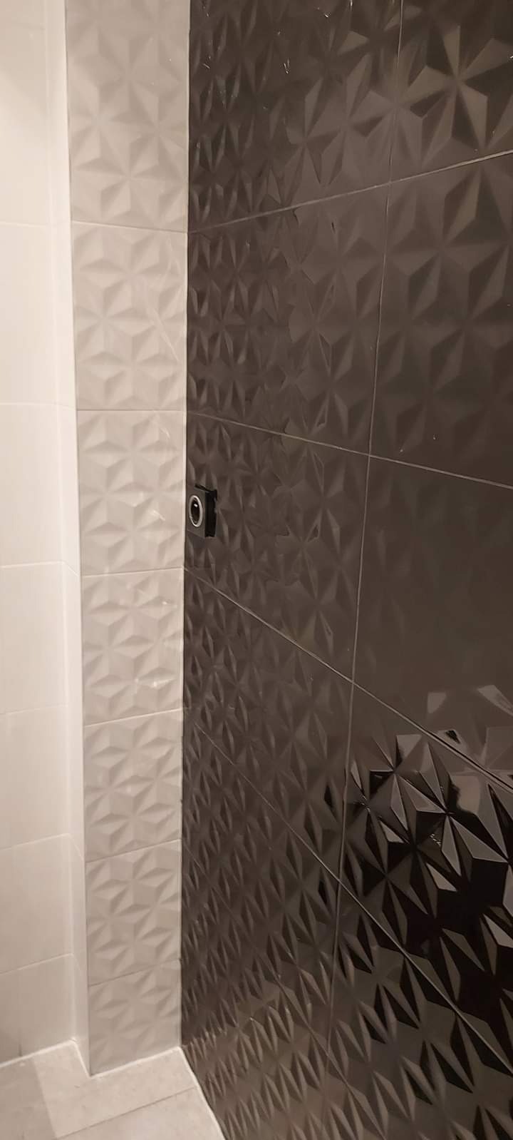 Ściana łazienki wyłożona białymi i grafitowymi płytkami z trójwymiarowym wzorem, widoczny fragment instalacji elektrycznej.