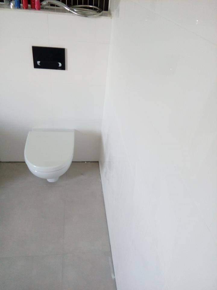 Nowoczesna toaleta podwieszana z czarnym przyciskiem spłukującym na białych płytkach ściennych i szarych płytkach podłogowych.