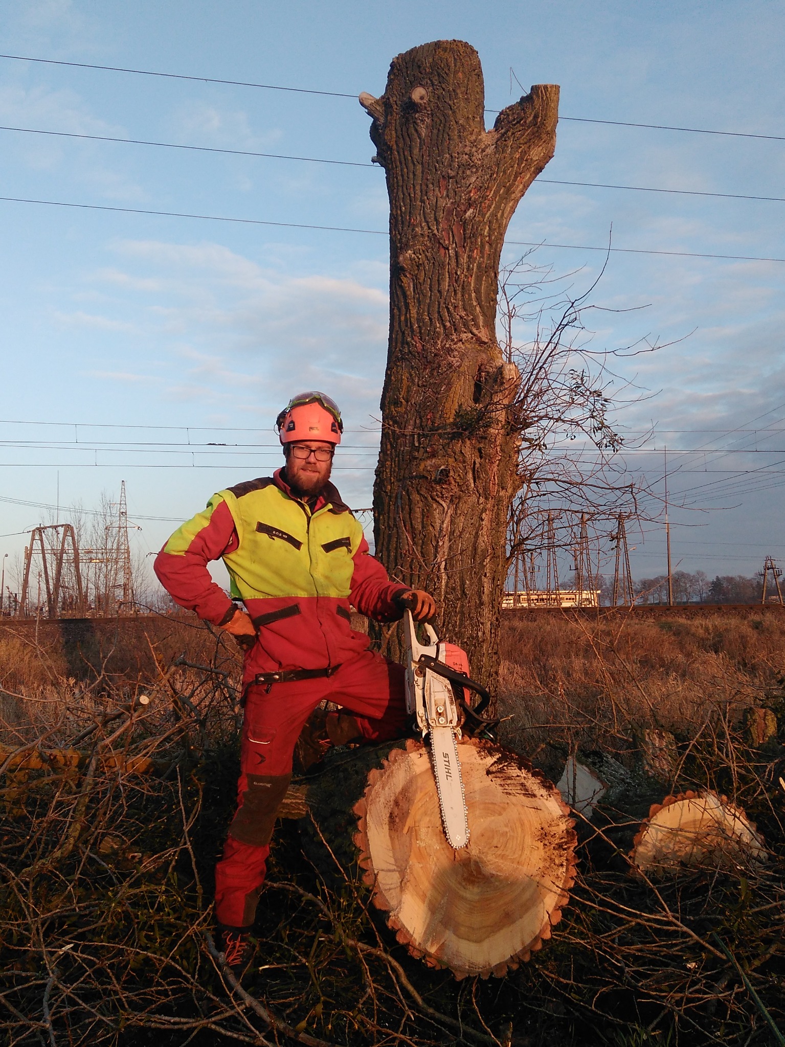 Pracownik w Otwocku, ubrany w odzież ochronną i kask, opiera się o ścięty pień drzewa z piłą spalinową Stihl, na tle linii kolejowej i słupów wysokiego napięcia.