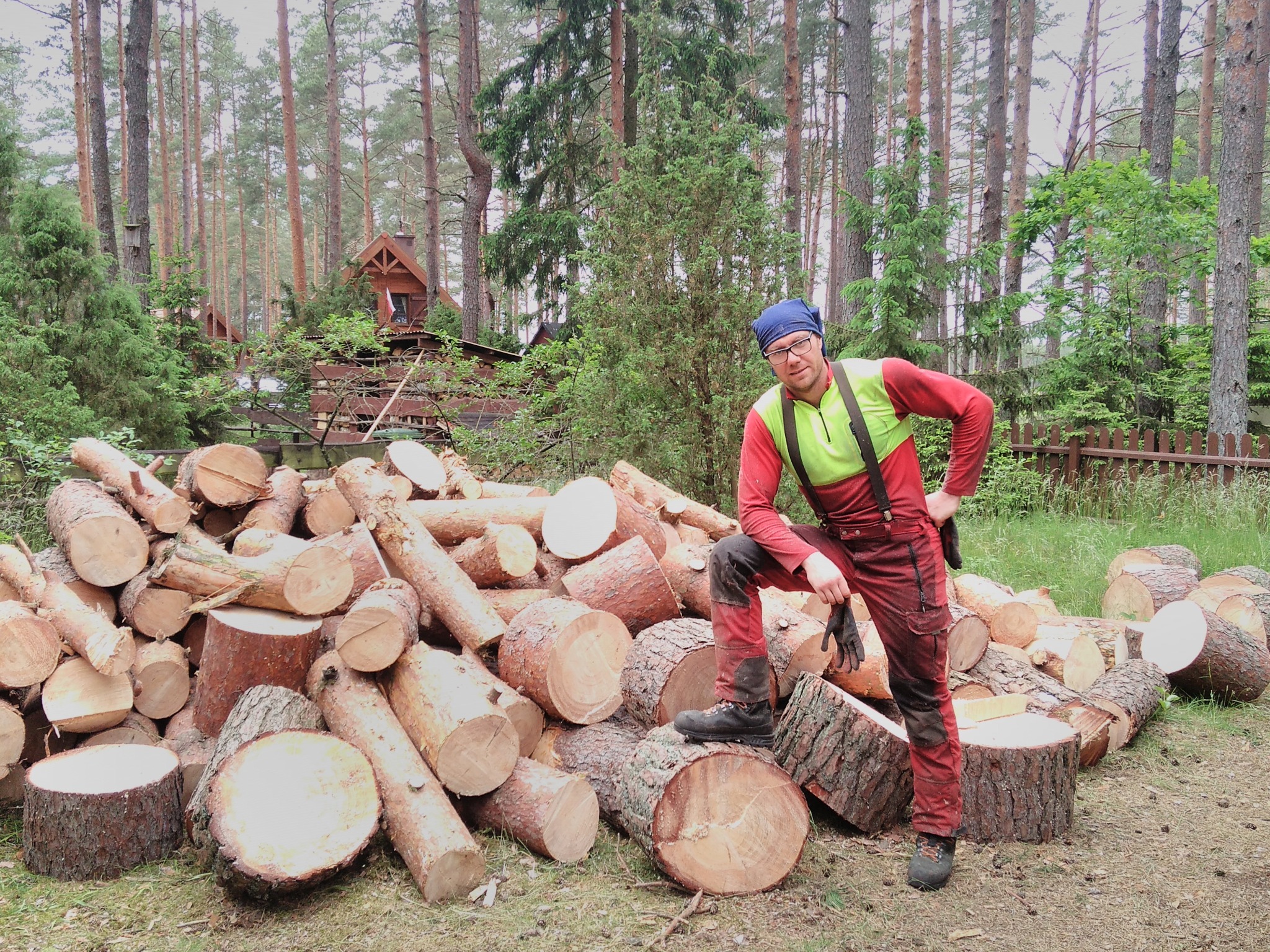 Mężczyzna w stroju roboczym opiera nogę na stosie pociętych pni drzew na tle leśnej chaty i drzew.