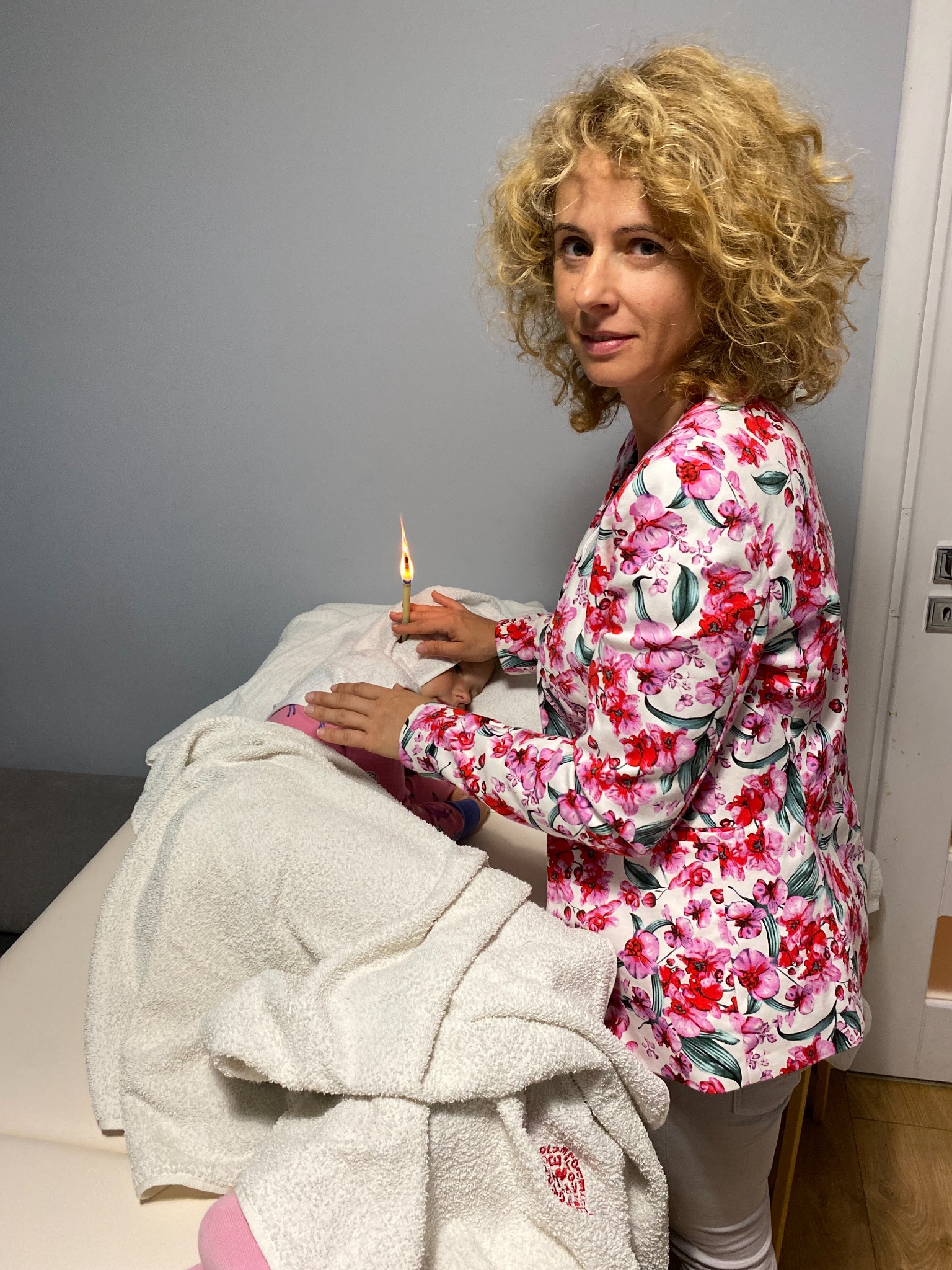 Kobieta w kwiecistej bluzce przeprowadza zabieg świecowania uszu u dziecka leżącego na leżance przykrytego ręcznikiem.