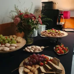 Stół cateringowy z ciastkami, owocami (truskawki, winogrona) i wafle na złotych talerzach, w tle ekspres do kawy i bukiet kwiatów.