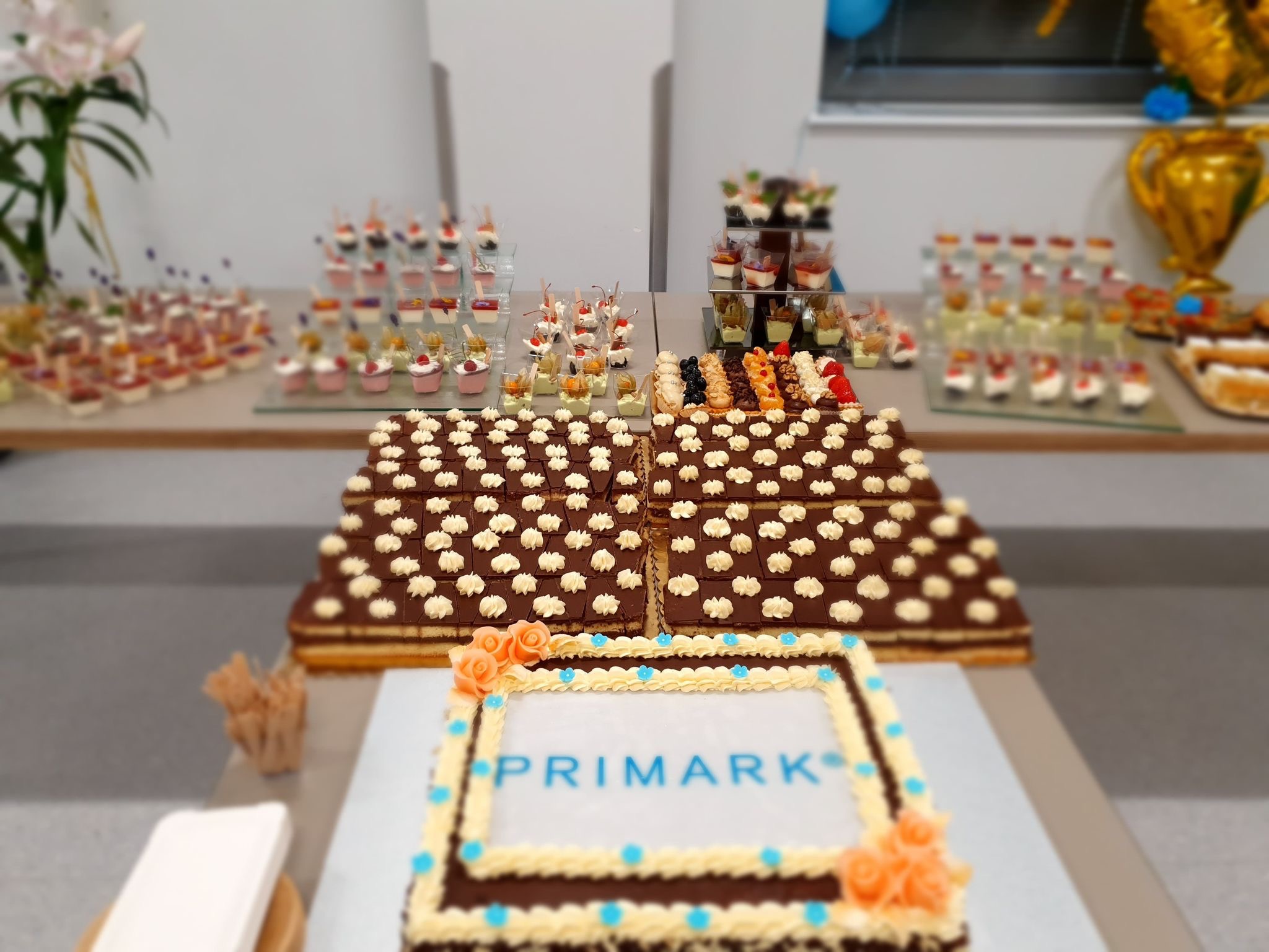 Stół z deserami na przyjęciu firmowym: tort z logo Primark, ciastka z kremem i mini desery w szklankach, złoty balon w kształcie pucharu.