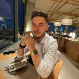 Młody mężczyzna z brodą, w koszuli w drobny wz&oacute;r, siedzi przy stole w restauracji z widokiem na miasto nocą; na stole leżą sztućce, telefon i butelki z napojami.