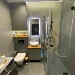 Nowoczesna łazienka z szarymi płytkami, kabiną prysznicową typu walk-in, umywalką nablatową na drewnianym blacie, podświetlanym lustrem i pralką z przodu.