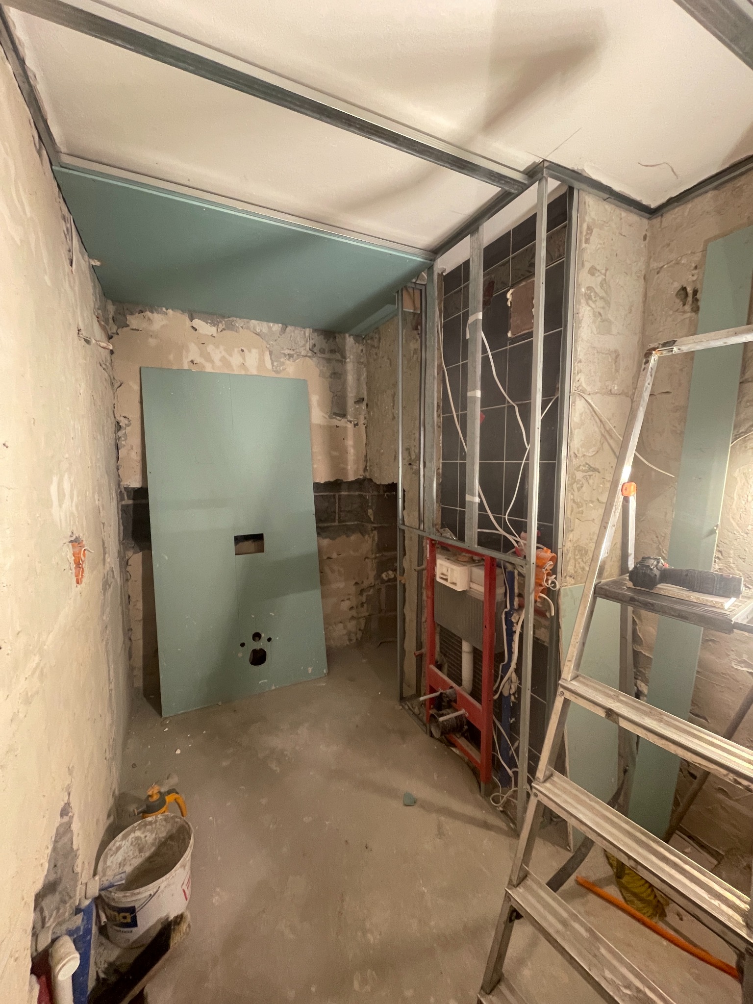 Wnętrze w trakcie prac: surowe ściany, metalowa konstrukcja pod zabudowę, płyta gipsowa oparta o ścianę, drabina aluminiowa i wiadro z resztkami zaprawy.