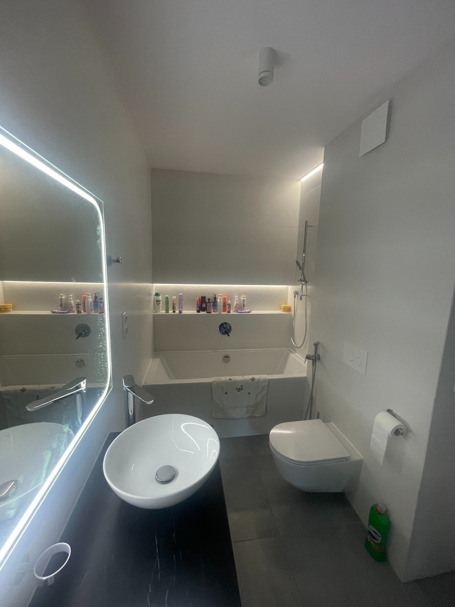 Nowoczesna łazienka w odcieniach szarości i bieli z podświetlanym lustrem, owalną umywalką nablatową na czarnym blacie, wanną z półką na kosmetyki i wiszącą toaletą.