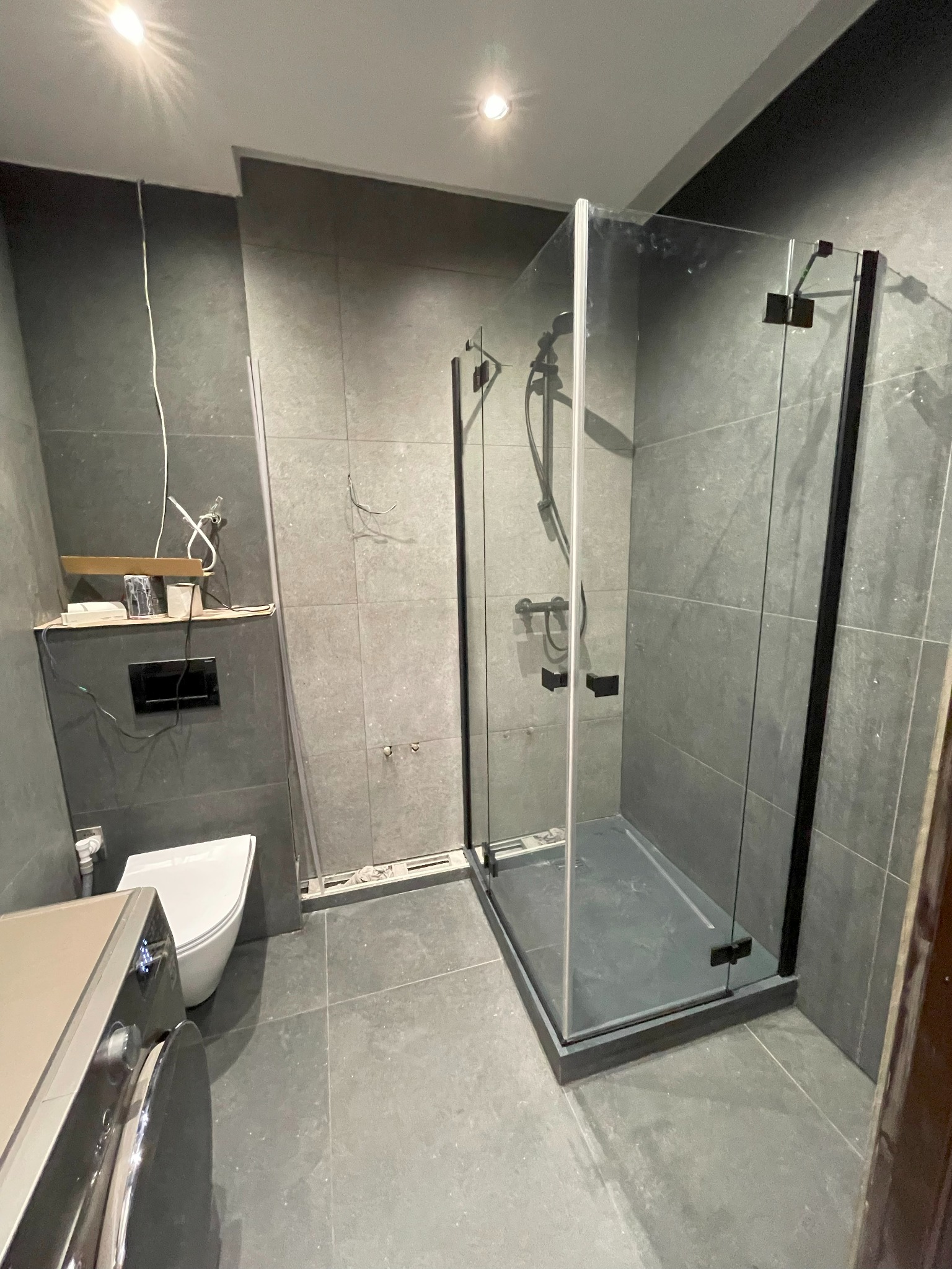 Szara łazienka w trakcie wykańczania: kabina prysznicowa z czarnymi ramami, podwieszana toaleta, pralka oraz niedokończone podłączenia elektryczne i hydrauliczne.