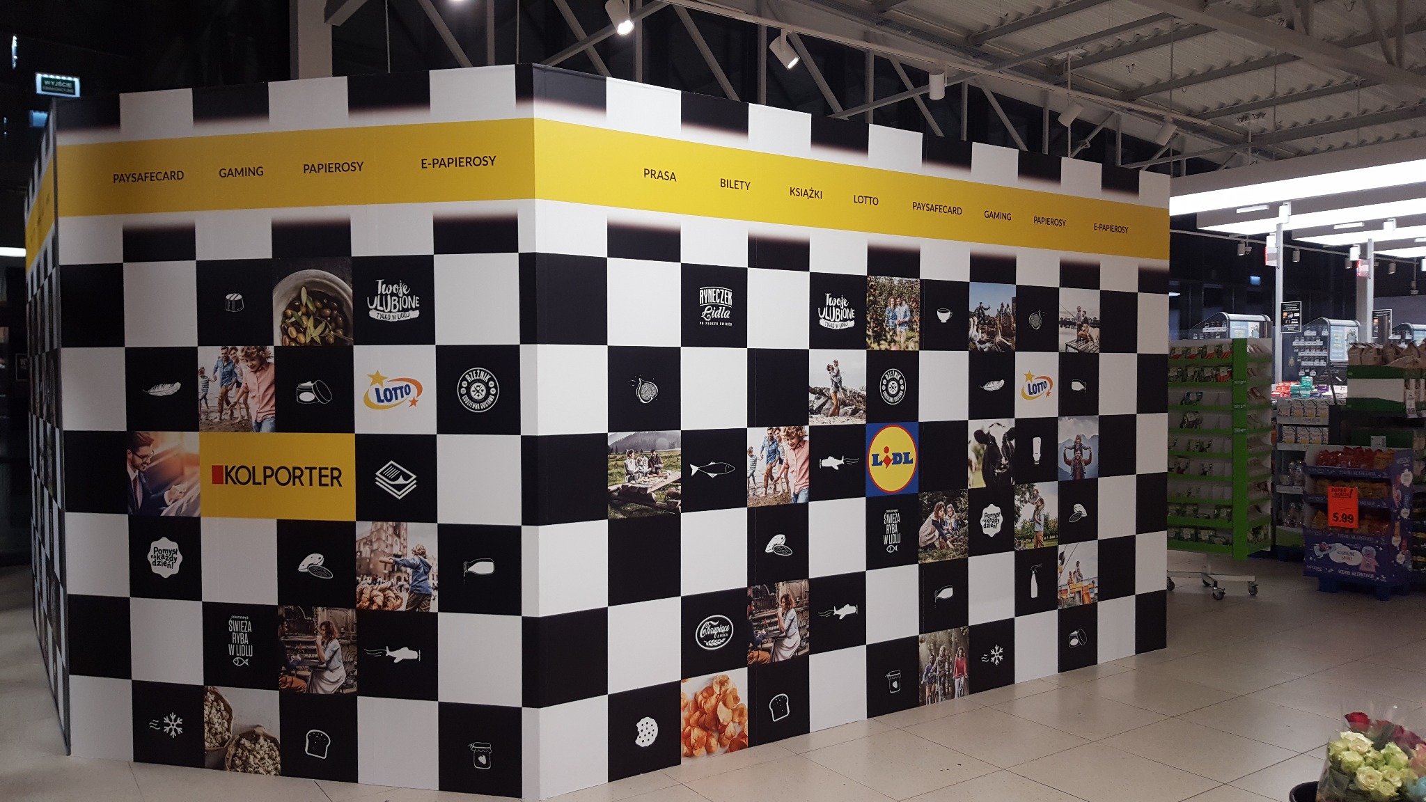 Stoisko promocyjne w supermarkecie z grafiką w szachownicę prezentującą logotypy i zdjęcia produktów, w tym Lotto, Kolporter, Lidl, oraz hasła reklamowe, z żółtymi nagłówkami kategorii produktów...