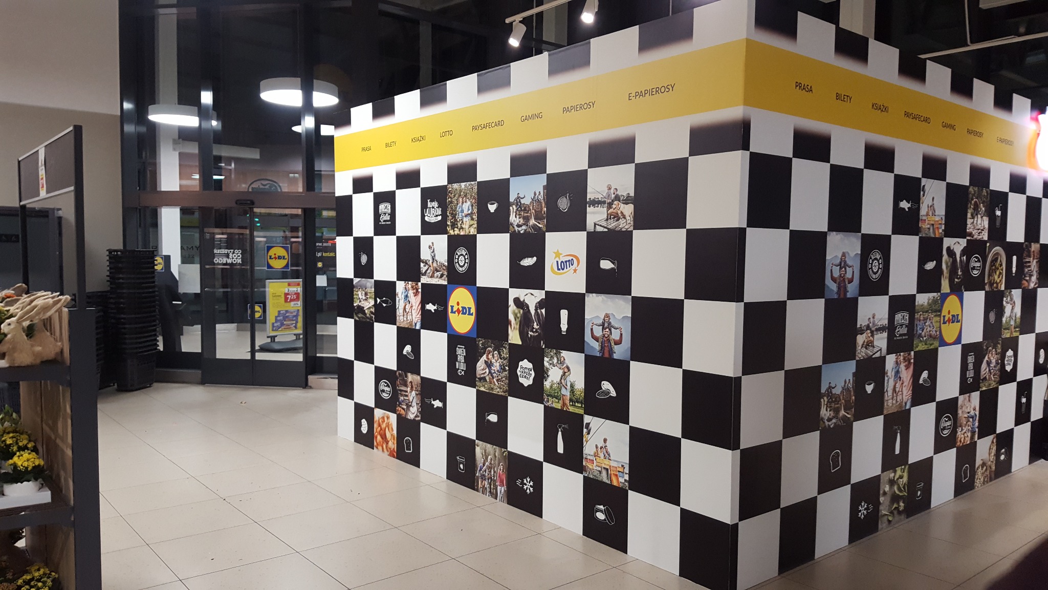 Ściana w markecie wyklejona czarno-białą szachownicą z wkomponowanymi zdjęciami i logotypami, nad nią żółty pas z napisami kategorii produktów, widoczne wejście do sklepu i koszyki na zakupy.