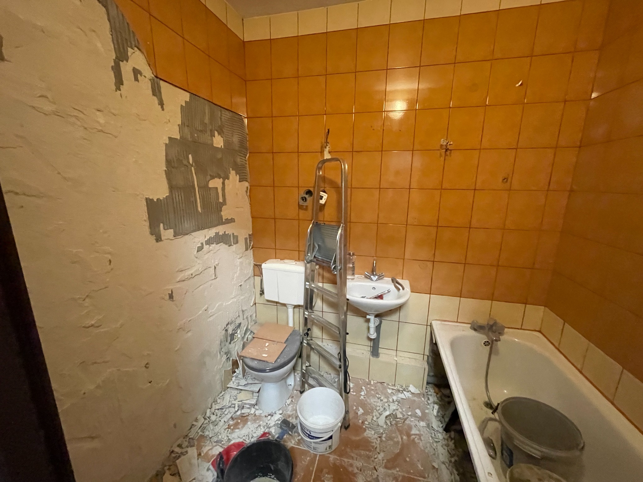 Demontaż starej łazienki: ściany z częściowo usuniętymi pomarańczowymi płytkami, gruz na podłodze, drabina oparta o umywalkę, toaleta z kartonami na desce, widoczne wiadra i narzędzia.