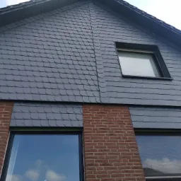 Fragment elewacji domu z szarym łupkiem i czerwoną cegłą, widoczne okna z odbiciem nieba.