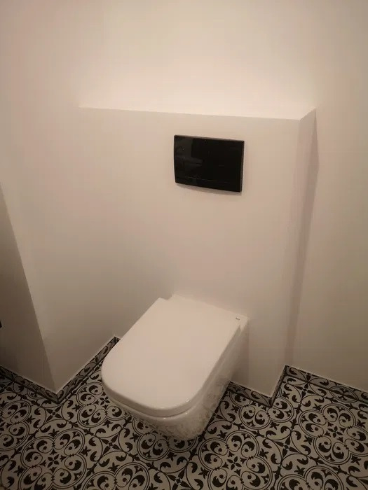 Wisząca toaleta z czarnym przyciskiem spłukującym na tle białej ściany z obudową i wzorzystą, czarno-białą podłogą.