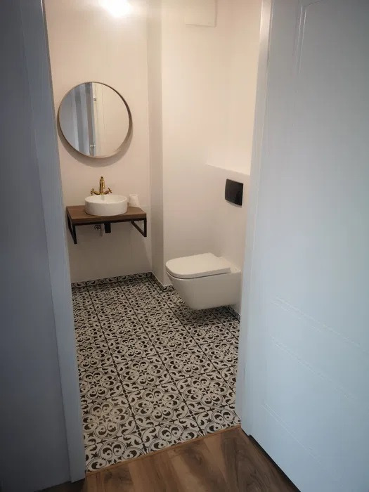 Mała łazienka z białą wiszącą toaletą, okrągłym lustrem nad umywalką z baterią w kolorze złota, oraz wzorzystą czarno-białą podłogą, widoczna zza uchylonych białych drzwi.