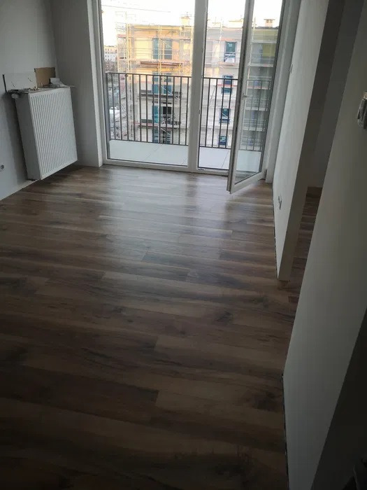 Nowe panele podłogowe w odcieniach brązu w jasnym pokoju z balkonem i widokiem na budowę za oknem.