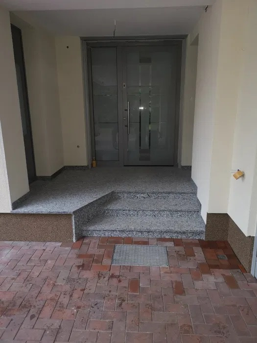 Wejście do budynku z szarymi, przeszklonymi drzwiami, schodami z lastryko i ceglaną posadzką. Widoczna kratka ściekowa w posadzce oraz cokół z imitacji kamienia na ścianach.