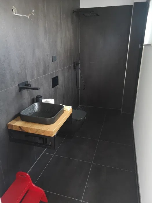 Nowoczesna łazienka z czarnymi płytkami, wiszącą umywalką z drewnianym blatem i czarną armaturą, widoczny czerwony stołek dla dziecka.