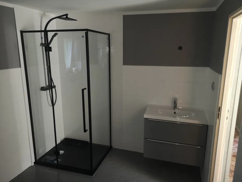 Nowoczesna łazienka z kabiną prysznicową typu walk-in z czarną ramą, szarą szafką pod umywalką i płytkami w odcieniach szarości i bieli.