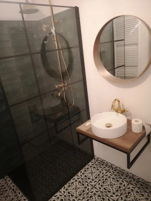 Nowoczesna łazienka z kabiną prysznicową typu walk-in z czarnymi profilami, okrągłym lustrem w złotej ramie, białą umywalką nablatową na drewnianym blacie z czarną metalową podstawą i wzorzystą...