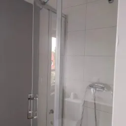 Szklana kabina prysznicowa z chromowanymi detalami, widoczny fragment toalety i armatura, świeżo po montażu, zabezpieczona niebieską taśmą malarską.