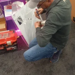 Elektryk w czapce '1932' klęczy na podłodze z wykładziną, podłączając przewody do panelu LED za pomocą śrubokręta, obok czerwony kufer narzędziowy.