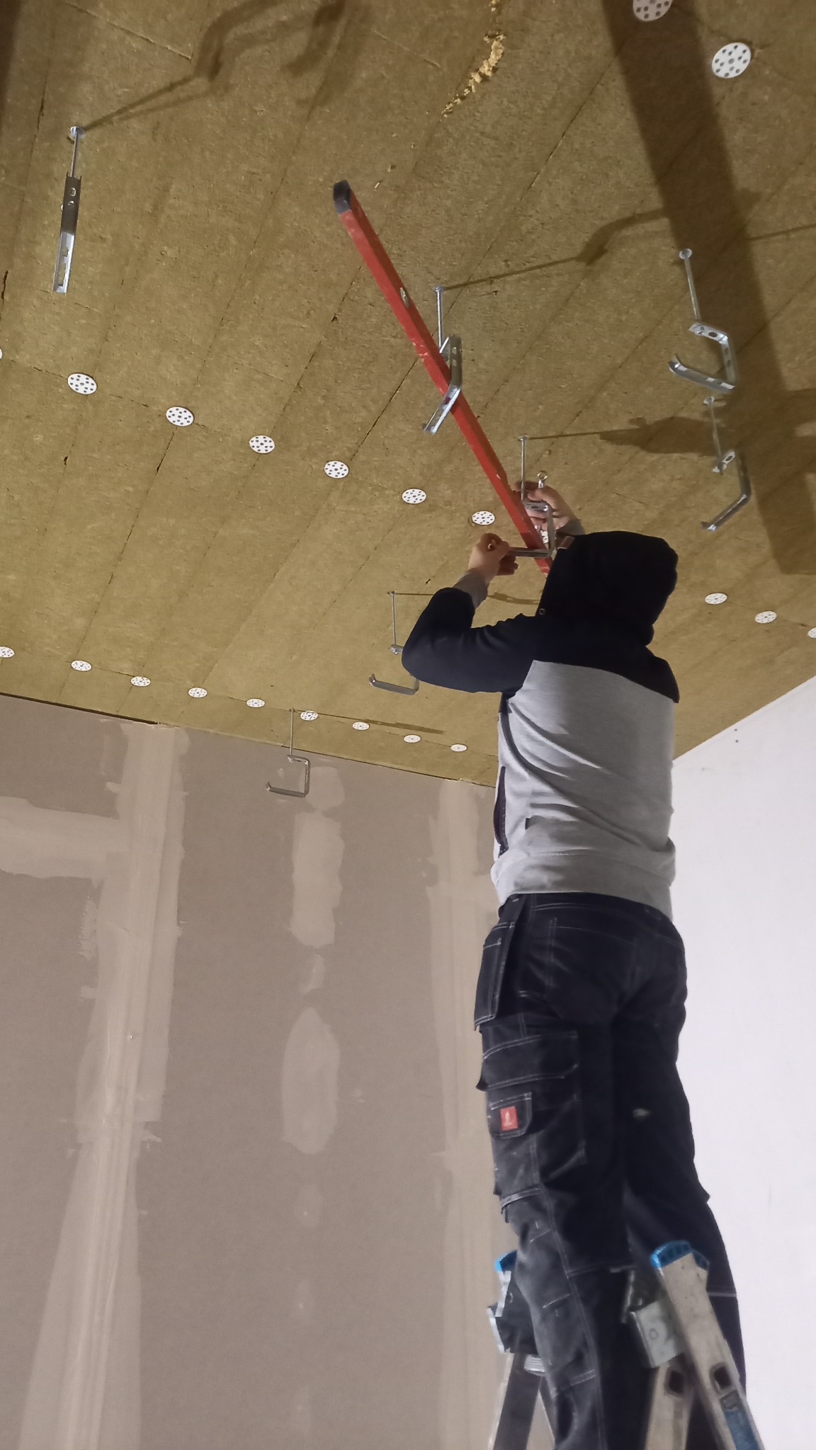 Pracownik na drabinie montuje konstrukcję podwieszanego sufitu z izolacją z wełny mineralnej, widoczne mocowania i profil.