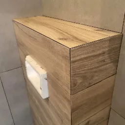 Zabudowa geberitu wykończona płytkami imitującymi drewno, z widoczną białą ramką wokół otworu rewizyjnego.