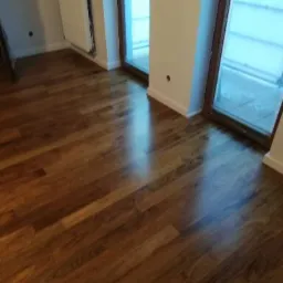 Błyszcząca, brązowa podłoga z desek ułożona w jasnym pomieszczeniu z grzejnikiem i oknami balkonowymi.