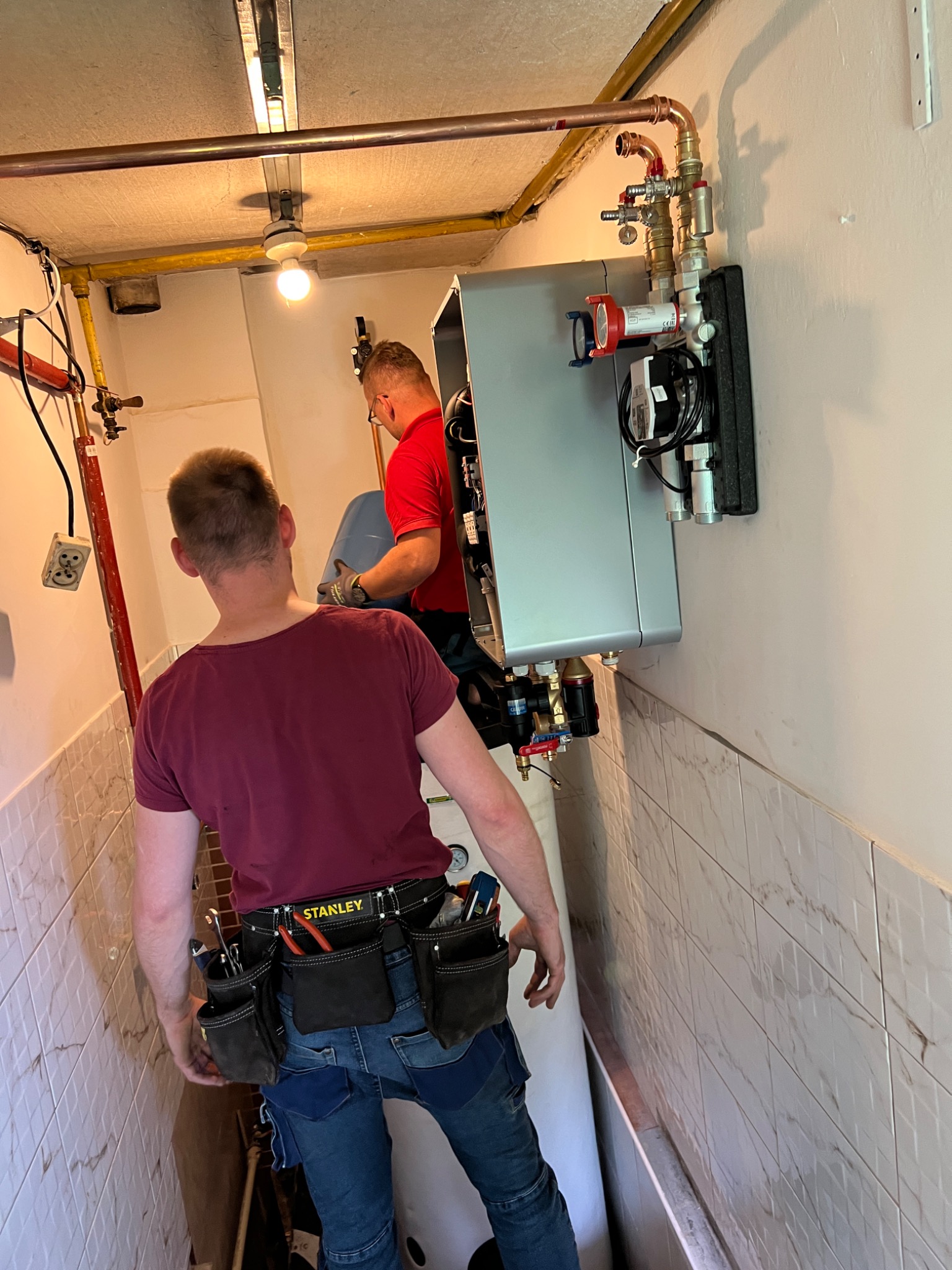 Dwóch instalatorów w trakcie montażu nowoczesnego pieca grzewczego w pomieszczeniu z wykończeniem imitującym marmur, widoczne miedziane rury i zawory, jeden z instalatorów ma na sobie pas...