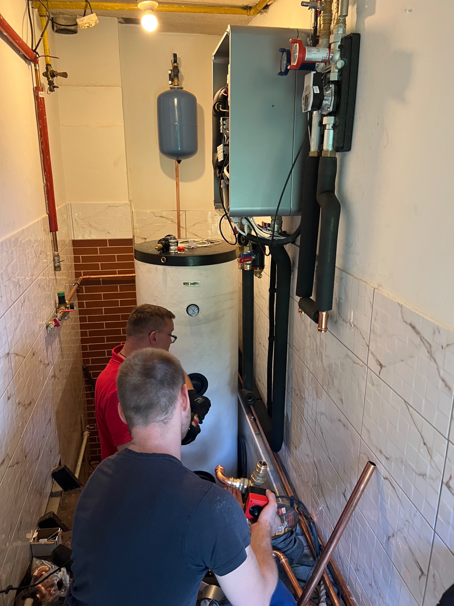 Instalatorzy w trakcie montażu kotła grzewczego w pomieszczeniu z marmurkowymi płytkami, widoczne liczne rury miedziane i osprzęt instalacyjny.