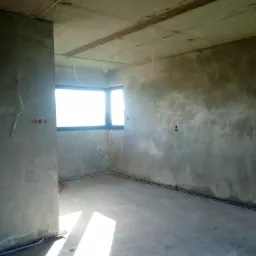 Surowe wnętrze w stanie deweloperskim, ściany pokryte świeżym tynkiem, widoczne przewody elektryczne, okno wpuszczające jasne światło słoneczne na podłogę.