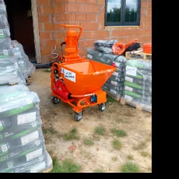 Pomarańczowa maszyna do tynkowania MAREK M-4 MAX na kółkach, obok palety z workami tynku maszynowego OZ421GT, widoczna ściana z pustaków ceramicznych i okno z roletą zewnętrzną.