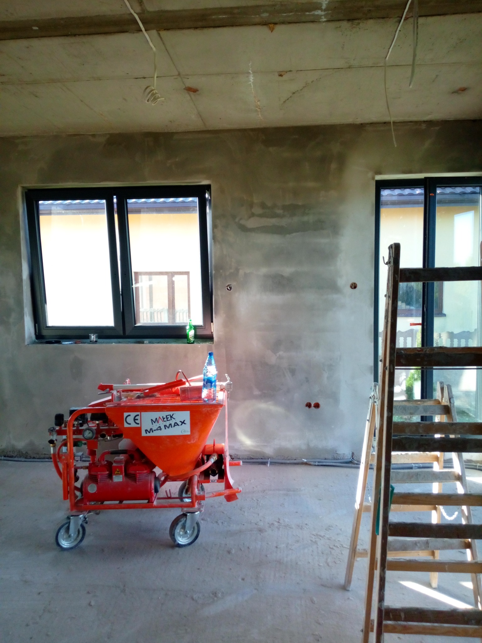 Wnętrze w trakcie tynkowania maszynowego: pomarańczowy agregat tynkarski Małek M-4 MAX, drewniana drabina i surowe ściany z widocznymi przewodami elektrycznymi.