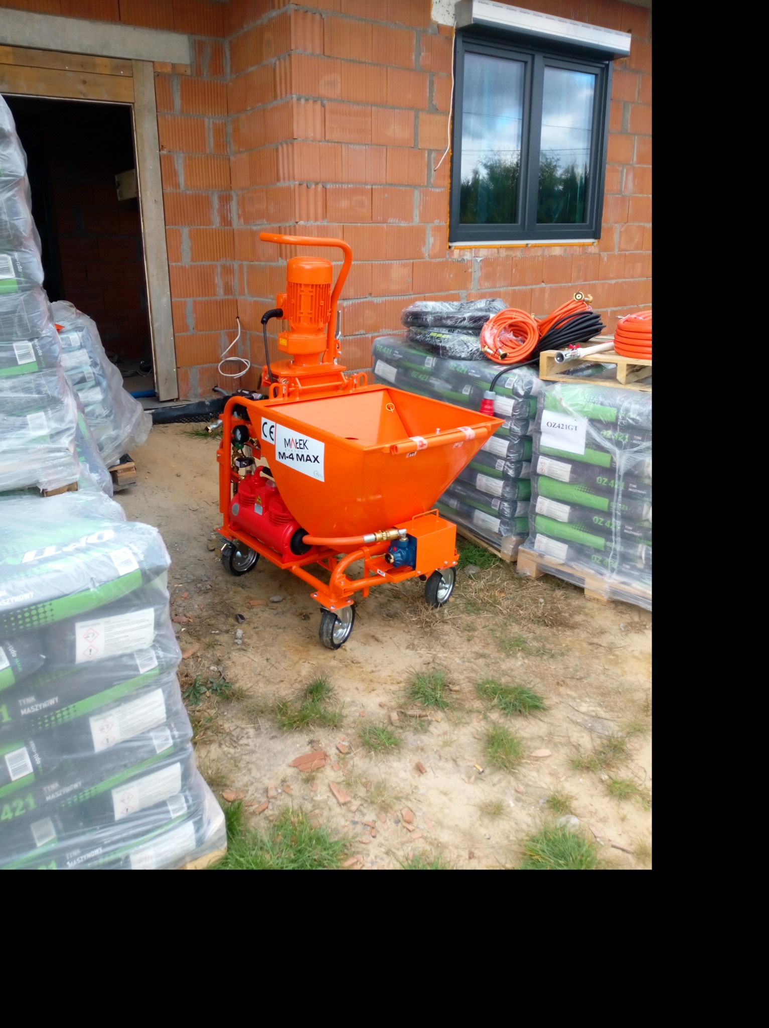 Pomarańczowa maszyna do tynkowania MAREK M-4 MAX na kółkach, obok palety z workami tynku maszynowego OZ421GT, widoczna ściana z pustaków ceramicznych i okno z roletą zewnętrzną.