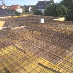 Zbrojenie stropu z siatki stalowej na żółtym deskowaniu, widoczne szalunki i betonowe bloczki, w tle zabudowa jednorodzinna.