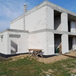 Surowy stan dwukondygnacyjnego domu murowanego z białych bloczków, widoczne otwory okienne i drzwiowe, częściowo otynkowany komin, drewniany kozioł murarski w pobliżu ściany, izolacja fundamentów.