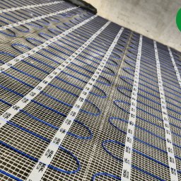 FOLIE GRZEWCZE, MATY -  Arkadiusz Zawadzki - Niebieskie przewody grzewcze ułożone na siatce z białymi taśmami mocującymi z nadrukiem 'HEAT DECOR' przed wylewką, widoczne logo producenta w prawym górnym rogu.