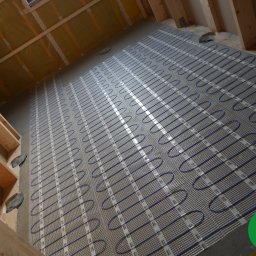 FOLIE GRZEWCZE, MATY -  Arkadiusz Zawadzki - Rozłożona mata grzewcza z niebieskimi przewodami na siatce, przygotowana do zalania wylewką, widoczne drewniane elementy konstrukcji stropu i ściany, w rogu logo producenta.