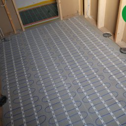 FOLIE GRZEWCZE, MATY -  Arkadiusz Zawadzki - Szara mata z niebieskimi kablami grzewczymi ułożona na podłodze w trakcie montażu, widoczne drewniane elementy konstrukcyjne ścian i logo producenta w prawym górnym rogu.