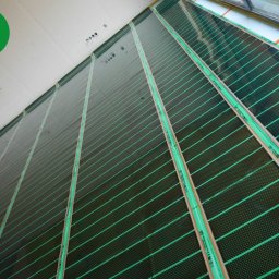 FOLIE GRZEWCZE, MATY -  Arkadiusz Zawadzki - Rozłożona, czarna mata z zielonymi paskami na podłodze, przygotowana do montażu ogrzewania podłogowego, widoczny fragment ściany i okna.