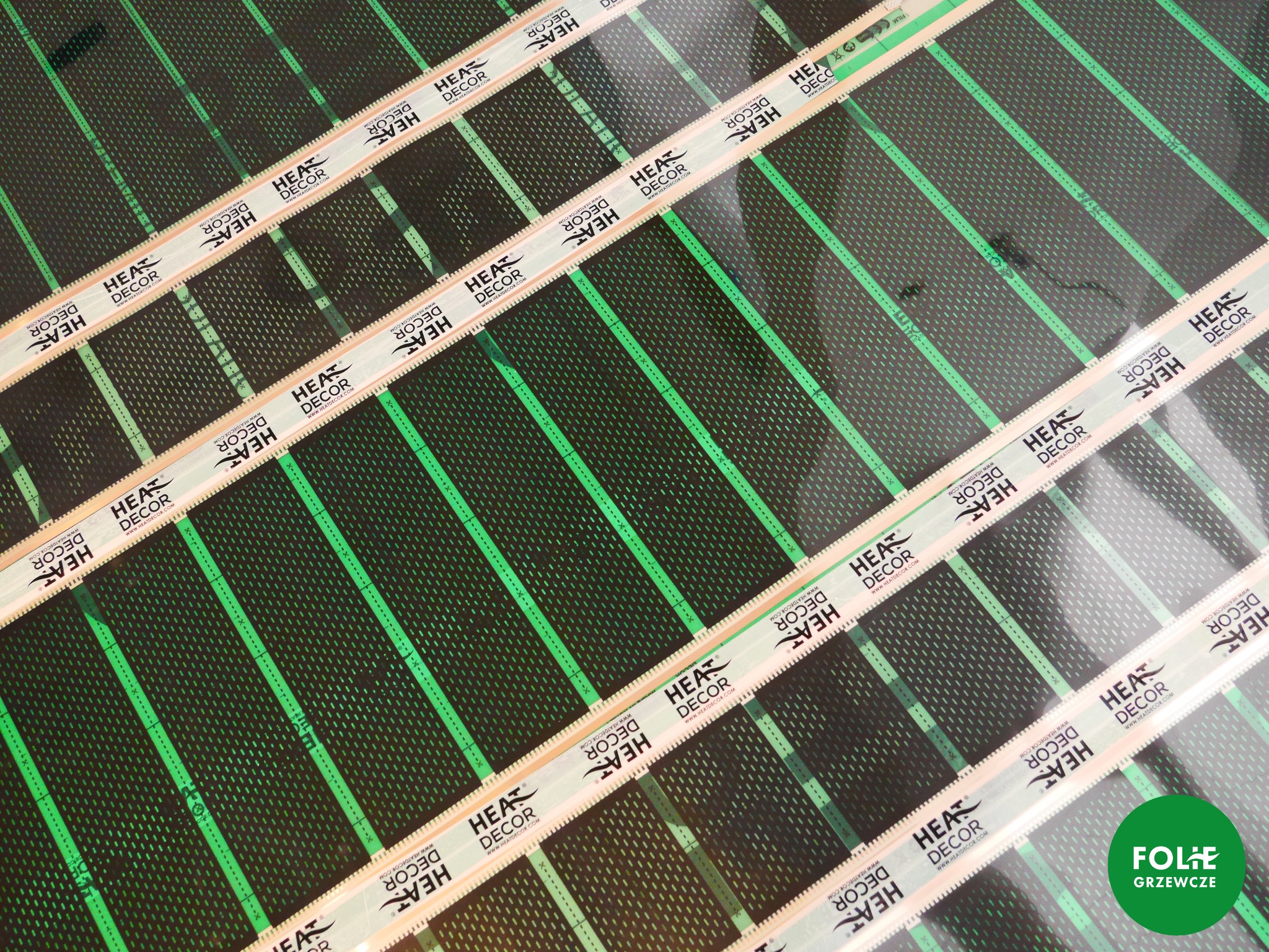 Fragment folii grzewczej z widocznymi paskami z nadrukowanym logo producenta Heat Decor, ułożonej na czarnym podłożu z zielonymi liniami.