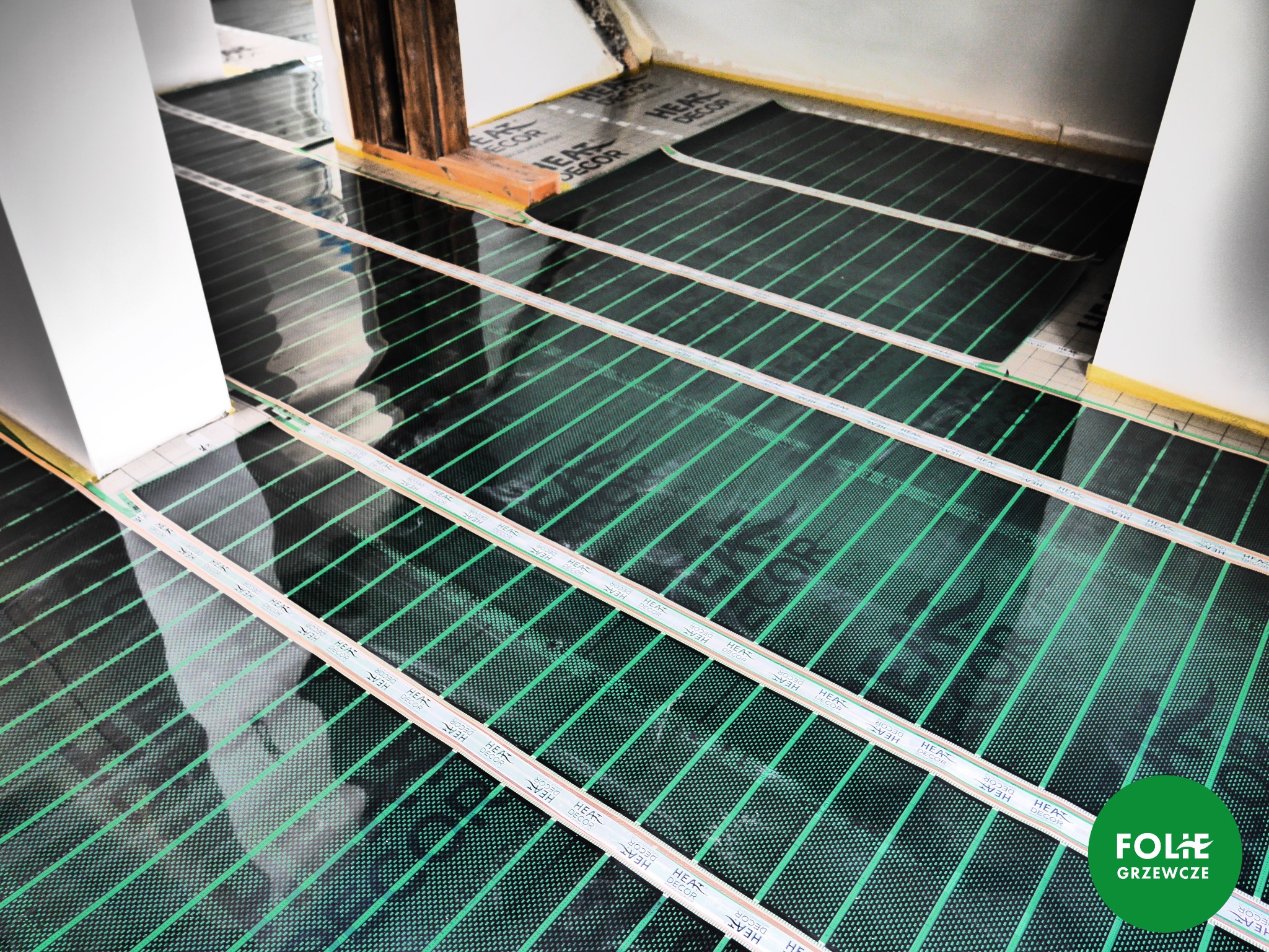 Rozłożone maty grzewcze z zielonymi paskami grzewczymi na czarnej folii, widoczne kable i taśmy montażowe, przygotowanie do montażu ogrzewania podłogowego w pomieszczeniu z surowymi ścianami.