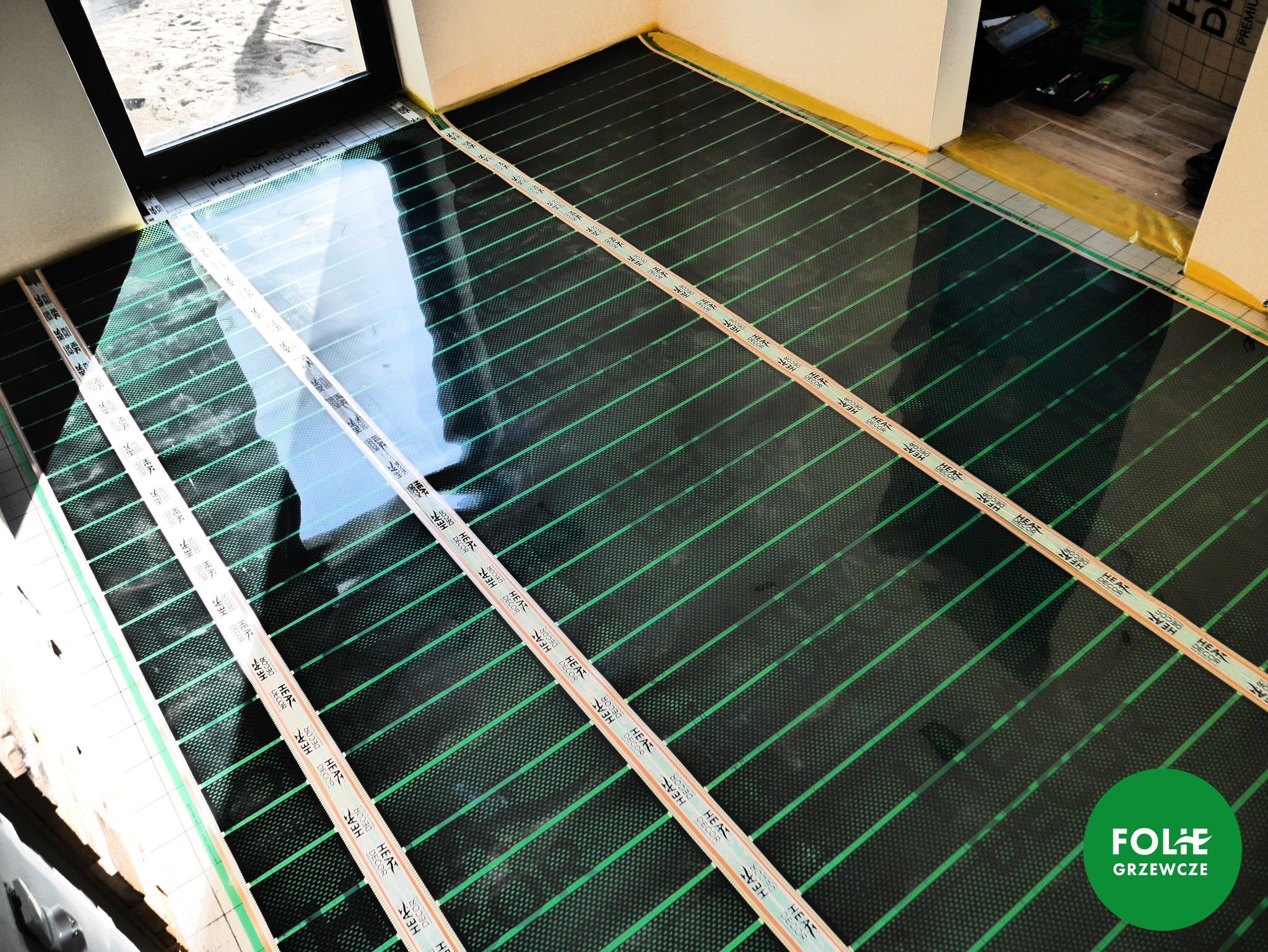 Rozłożona mata grzewcza z zielonymi liniami grzewczymi, zabezpieczona taśmą montażową z napisem 'HEAT DECOR', przed montażem podłogi, widoczne okno i przygotowane narzędzia w tle.