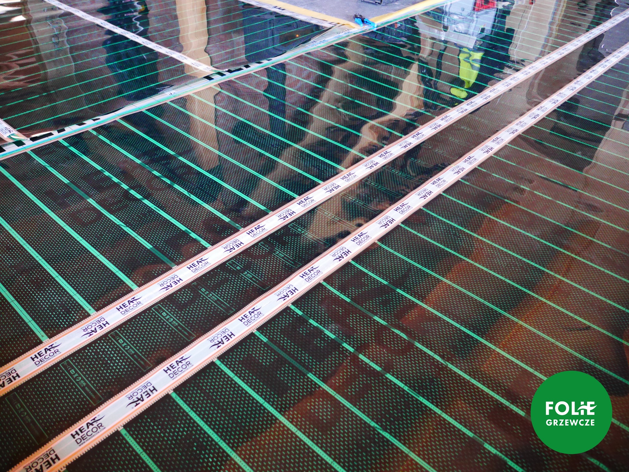 Rozłożona folia grzewcza z zielonymi liniami grzewczymi, zabezpieczona taśmą z logo 'HEAT DECOR', widoczne odbicie osoby w tle.